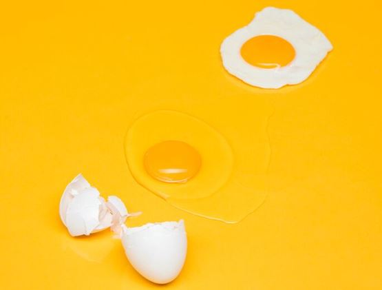 A cracked egg, a raw yolk, and a fried egg arranged on a bright yellow background.