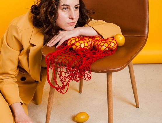 A woman wearing a mustard-yellow suit leans on a brown chair with lemons placed in a red net bag, against a yellow background.
