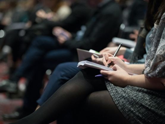 People sitting in a conference audience taking notes in notebooks during a presentation or seminar.