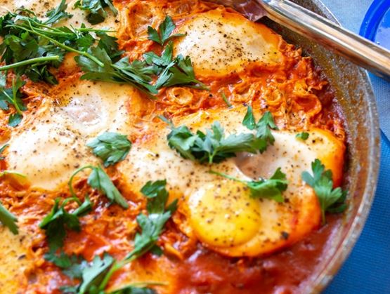 A close-up of shakshuka in a pan, with poached eggs in tomato sauce topped with fresh parsley and served with bread and cheese on the side.