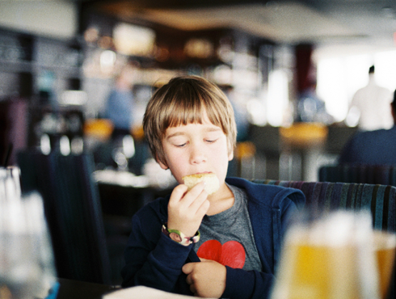 Child sitting in a restaurant, eating a piece of bread while looking down thoughtfully.