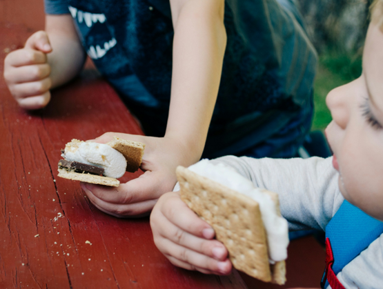 Children holding s’mores with melted marshmallows and chocolate at an outdoor table.