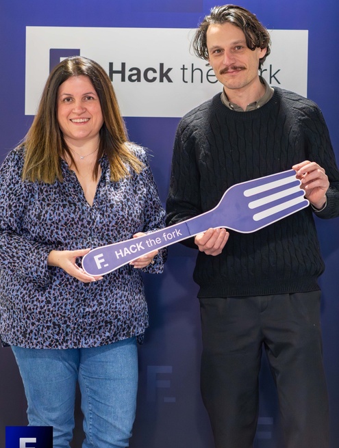 Two people pose in front of a “Hack the fork” backdrop holding a large purple fork with the event logo.