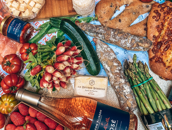 Food spread still life with tomatoes, radishes, zucchini, asparagus, cured sausage, bread, cheese, jam, strawberries and a bottle of rosé on a table.