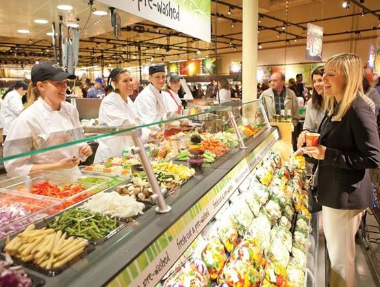 Supermarket salad bar counter with staff in uniforms serving customers in front of a vegetable display.