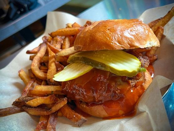 Sauce-covered burger topped with pickle slices, served with fries in a paper-lined tray.