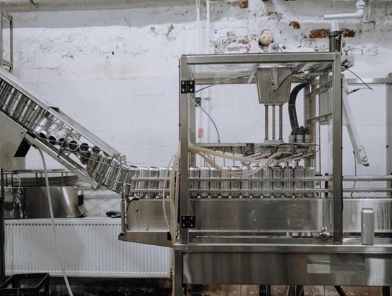 Stainless-steel industrial machine in a factory, with a conveyor line and rows of containers lined up.