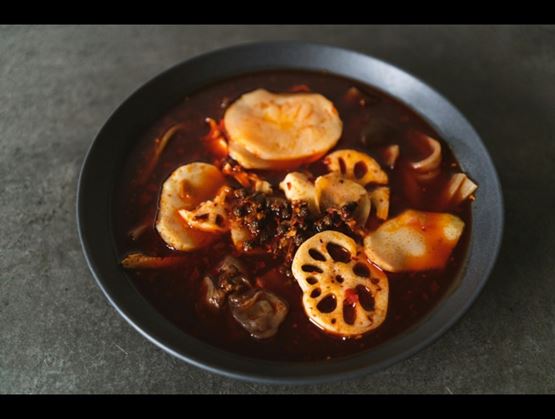 Bowl of spicy red soup with sliced lotus root and other ingredients, served in a black bowl.