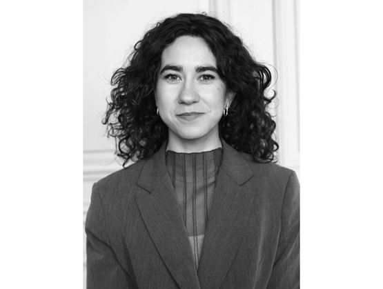 Black and white portrait of a woman with curly brown hair, wearing earrings and a dark blazer with a sheer top, standing against a light indoor background.