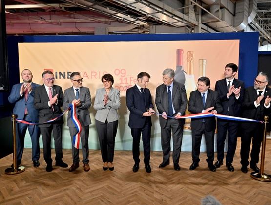 Official inauguration ceremony with several dignitaries standing in a row cutting a tricolor ribbon on an indoor stage, in front of a display panel featuring bottles, while others applaud.