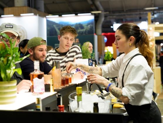Mixology demonstration at a trade show booth: a bartender prepares cocktails in front of attentive visitors, with bottles and glasses arranged on the counter.