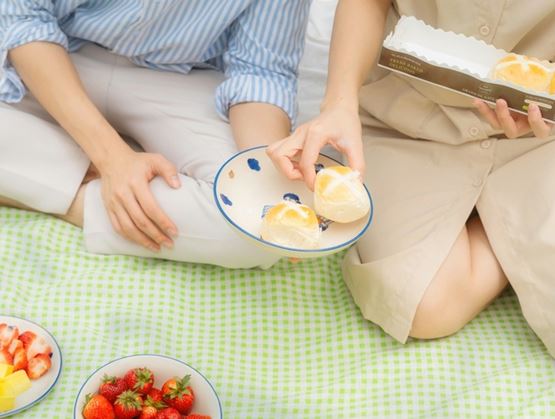 Two people sitting on a checkered blanket sharing bread rolls during a picnic, with fresh fruit arranged on plates.