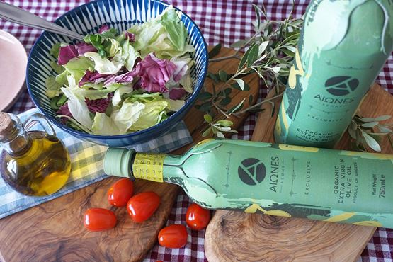 Bouteilles d’huile d’olive Aiones avec salade, tomates cerises et huilier sur table à carreaux.