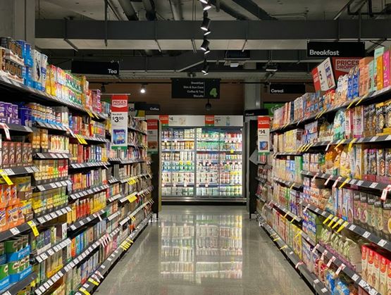Supermarket aisle with shelves stocked with food products and cereals on both sides, leading to refrigerated cases at the back under modern lighting.