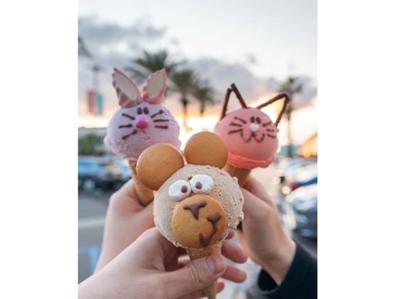 Three animal-shaped ice cream cones (rabbit, bear, and cat) held in hands outdoors, with palm trees and a sunset sky in the background.
