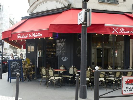 Façade d’un bistrot parisien avec auvent rouge portant l’inscription « Bistrot le Peletier », terrasse extérieure équipée de tables et chaises en rotin, ardoise de menu et rue urbaine en arrière-plan.