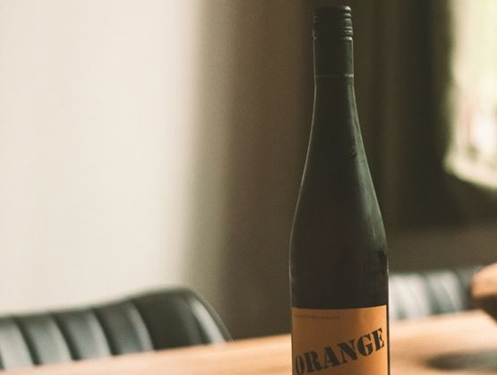 Bouteille de vin orange posée sur une table en bois dans un intérieur tamisé, avec une étiquette minimaliste et une lumière naturelle venant d’une fenêtre en arrière-plan.