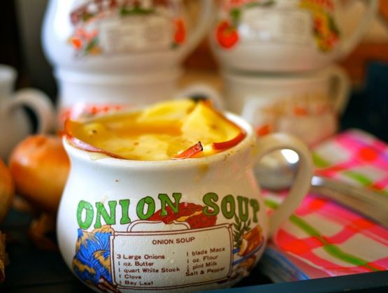 Bol de soupe à l’oignon gratinée servi dans une tasse décorée avec l’inscription « Onion Soup » et la recette imprimée, garni de fromage fondu et de tranches d’oignon, avec des oignons et des récipients en arrière-plan.