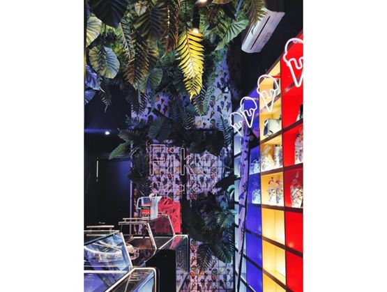 Intérieur coloré d’un salon de glaces avec décor tropical, plantes suspendues au plafond, vitrines réfrigérées et étagères lumineuses ornées de néons en forme de glace.