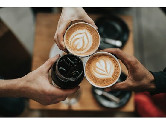 Three hands clink cups of coffee and a dark drink over a wooden table.