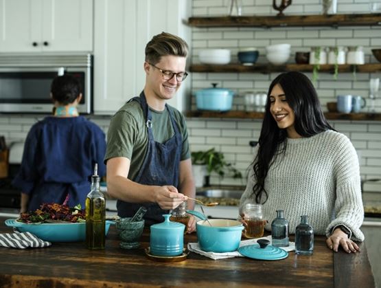 Deux personnes souriantes préparent un plat dans une cuisine moderne, en ajoutant un ingrédient dans une casserole posée sur un îlot en bois, avec des étagères et ustensiles en arrière-plan.
