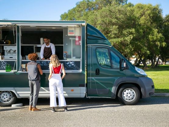 Deux personnes commandent de la nourriture à un food truck vert stationné dans un parc, tandis que le vendeur prend leur commande au comptoir extérieur.