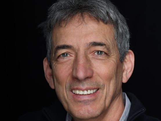 Professional portrait of a smiling man with short gray hair, wearing a light shirt and a dark jacket, against a black background.