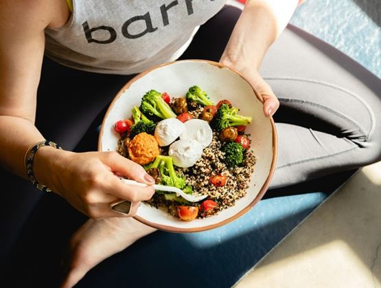 Une personne assise tenant un bol composé de quinoa, de brocoli, de tomates cerises et de fromage, utilisant une fourchette en plastique pour manger.