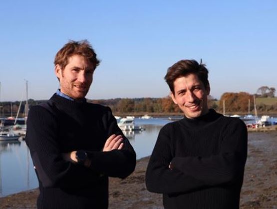 Deux hommes debout côte à côte près d’un port avec des bateaux en arrière-plan, les bras croisés et souriant à la caméra.