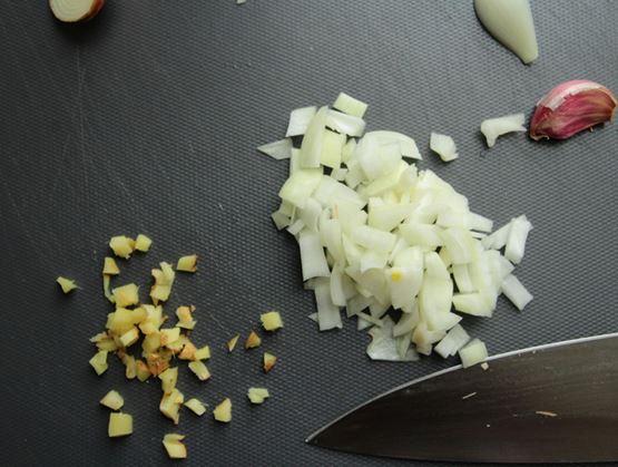 Chopped onion and minced ginger on a dark cutting board, with a garlic clove and a kitchen knife.