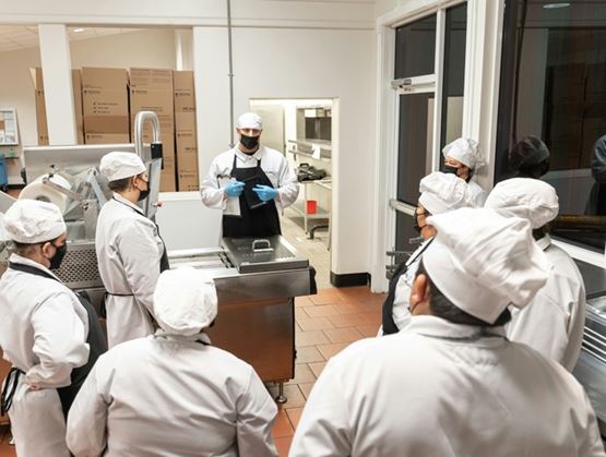 Une équipe de professionnels en tenue de cuisine et masques sanitaires réunie dans un atelier de transformation alimentaire, écoutant un responsable donnant des instructions autour d’un équipement industriel.