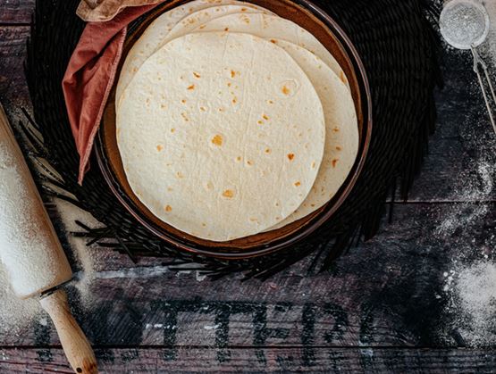 Stacked tortillas on a rustic plate, surrounded by flour, a rolling pin, and a sifter on a wooden table.