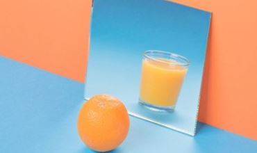 Orange entière posée devant un miroir reflétant un verre de jus d’orange, sur un fond coloré bleu et orange.