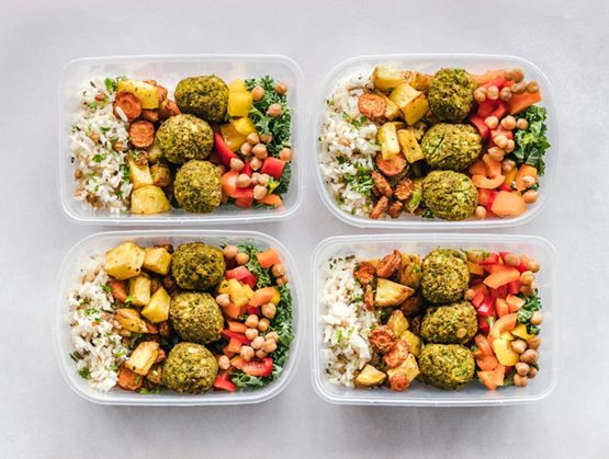 Four meal prep containers filled with rice, falafel balls, chickpeas, roasted vegetables, and colorful fresh vegetables arranged on a grey background.