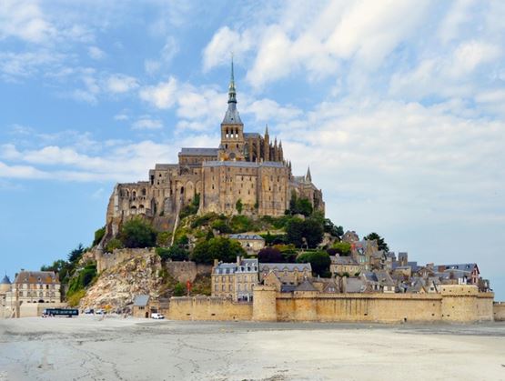Vue du Mont-Saint-Michel et de son abbaye perchée sur un îlot rocheux, entourée de remparts et de maisons, avec une vaste étendue de sable au premier plan.