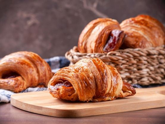 Viennoiseries feuilletées garnies, dorées et brillantes, présentées sur une planche en bois avec un panier en arrière-plan.
