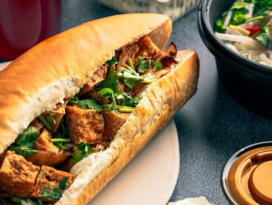 Sandwich vietnamien type bánh mì garni de viande et d’herbes fraîches, accompagné de rouleaux de printemps, d’une boisson et de plats asiatiques à emporter.