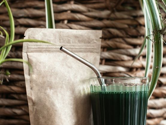 Verre de boisson verte à base de spiruline avec une paille en métal, posé à côté d’un sachet de poudre sur une table, entouré de plantes d’intérieur.