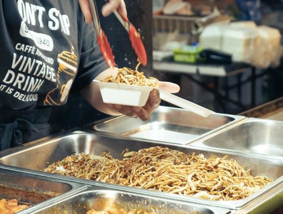 Personne utilisant une pince pour servir des nouilles sautées et des plats chauds depuis un buffet de street food dans une barquette.
