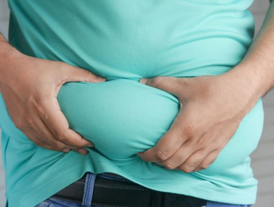 Personne pinçant la graisse de son ventre à travers un t-shirt turquoise, illustrant l’excès de graisse abdominale.
