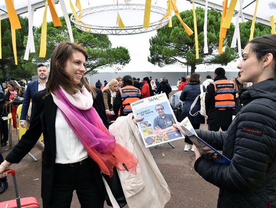 Scène d’entrée d’un événement en plein air où une hôtesse remet un programme à une visiteuse souriante, entourées de participants et de personnel de sécurité, sous une installation décorative avec rubans colorés.