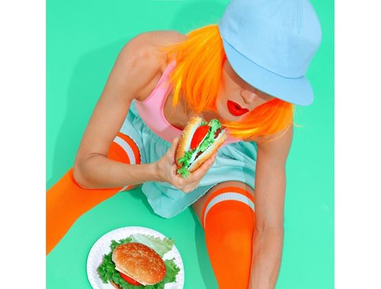Person with bright orange hair and a blue cap holding a burger, sitting on a turquoise background with another burger on a plate.
