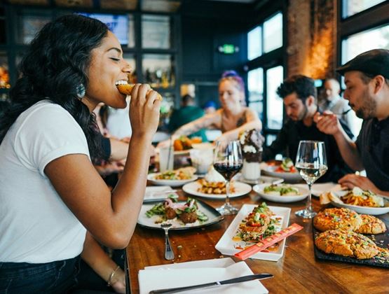 Groupe de personnes attablées dans un restaurant, dégustant différents plats et desserts ; au premier plan, une femme souriante mange, tandis que d’autres convives partagent des assiettes variées avec des verres de vin sur une table en bois.
