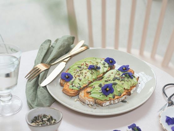 Assiette de tartines à l’avocat garnies de graines et de fleurs comestibles violettes, posée sur une table avec des couverts, un verre d’eau et une serviette, dans un cadre lumineux et élégant.