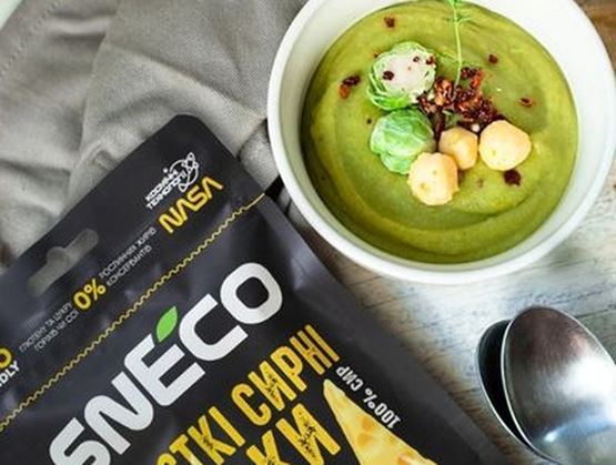 Bol de soupe verte garnie accompagné d’un snack au fromage innovant présenté dans son emballage, posé sur une table avec une cuillère.