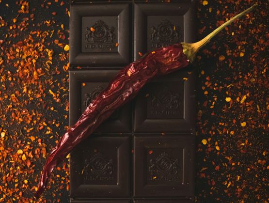 Tablette de chocolat noir posée sur un fond sombre, parsemée d’éclats d’épices et surmontée d’un piment rouge séché, évoquant une association gourmande et épicée.