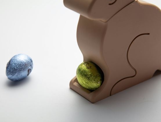 Stylized wooden or plastic bunny holding a shiny green egg, with another metallic blue egg placed nearby on a minimal neutral background.