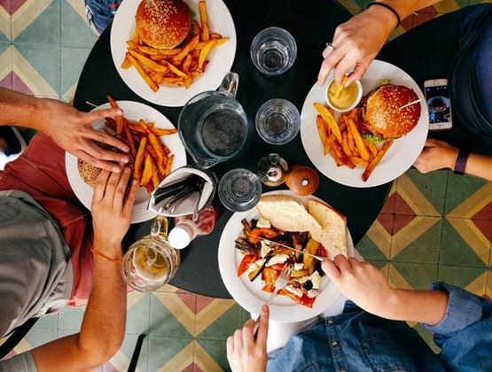 Vue de dessus d’une table de restaurant où plusieurs personnes partagent un repas : burgers accompagnés de frites, assiettes de légumes grillés avec pain, verres d’eau et bière, mains en train de manger et de tremper des frites dans une sauce.