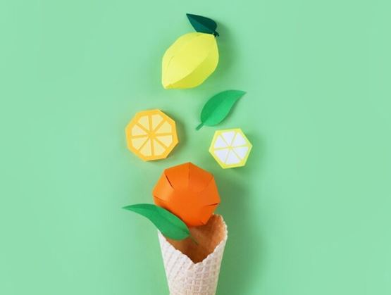 Colorful composition of stylized paper fruits, including a lemon and an orange, appearing to come out of an ice cream cone on a pastel green background, creating a playful and creative visual.