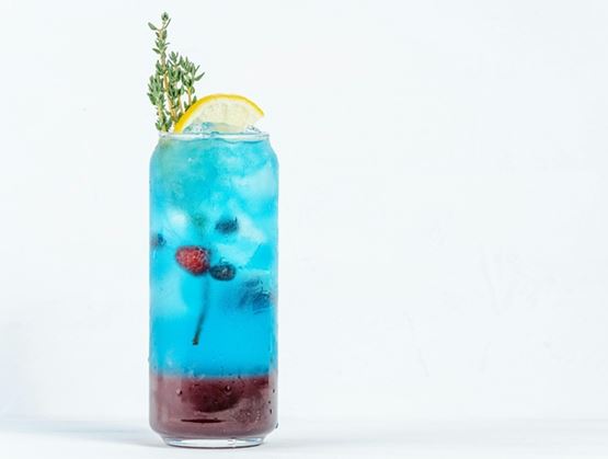 Refreshing blue drink served in a glass with ice, garnished with lemon, berries, and a sprig of herbs on a light background.